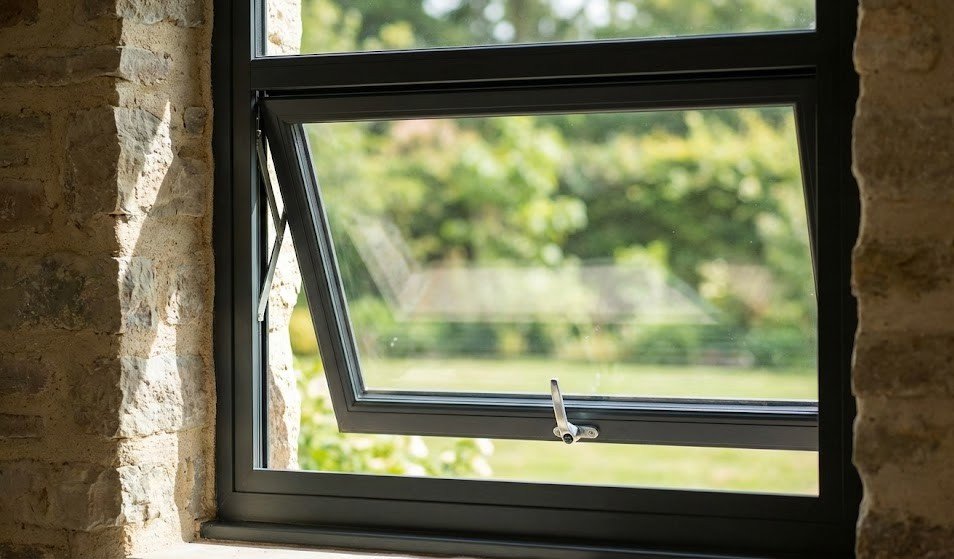 Modern black casement window open outward in a traditional stone wall, framing a sunny view of a lush green garden during a bright day.