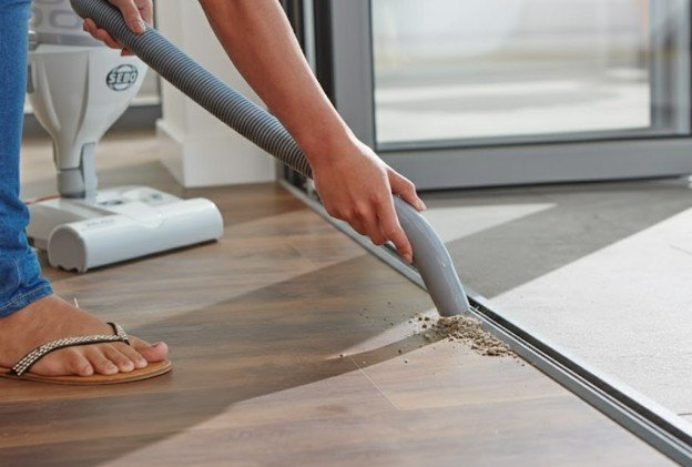 Person vacuuming dirt and debris along a sliding door track on a wooden floor using a hose attachment; only their lower legs and sandals are visible.